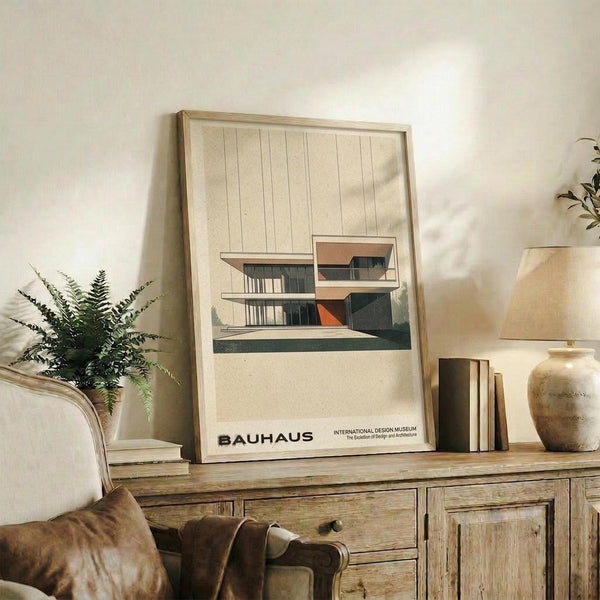 May include: Framed Bauhaus poster featuring modernist architectural illustration in muted beige, orange and green tones, displayed on rustic wooden console alongside fern plant, ceramic vases, and books in neutral living room setting with beige armchair and warm lighting