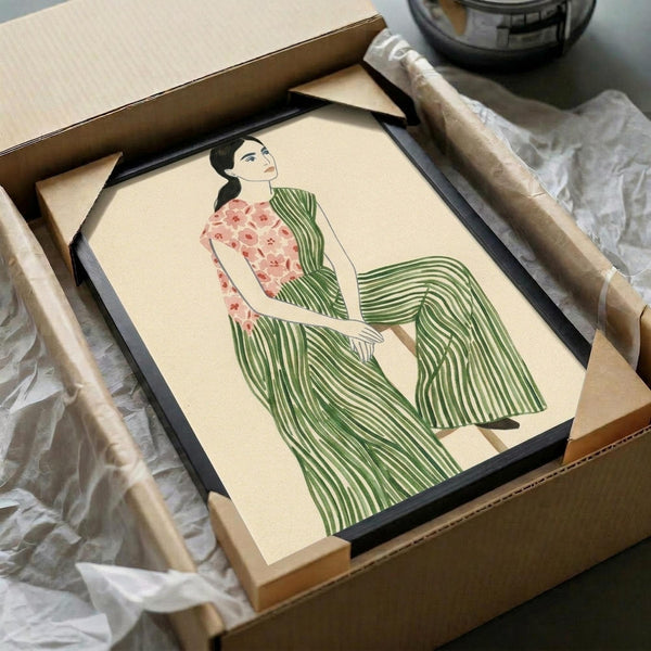 May include: Framed art print in black frame showing woman in green striped dress with floral pink top, sitting gracefully. Illustration has vintage-inspired style with cream background. Frame appears in cardboard shipping box with protective tissue paper wrapping. Wooden rolling pin visible beside packaging materials on white surface.