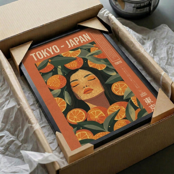 May include: Vintage-style poster with orange and green color scheme featuring stylized woman's face surrounded by citrus fruits and leaves, displayed in black frame inside cardboard shipping box with protective wrapping materials on white surface