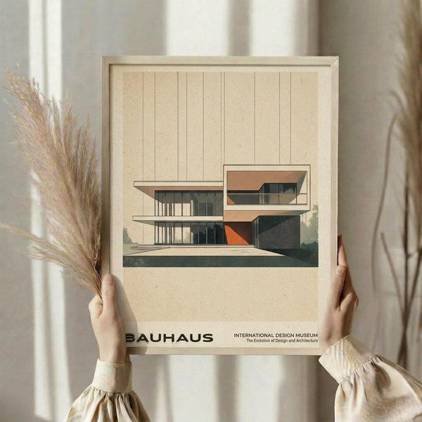 May include: Person holding framed Bauhaus poster featuring modernist architectural illustration of multi-level house with geometric design in beige, orange, and dark green colors. Cream-colored mat, natural wood frame. Soft lighting with dried pampas grass and neutral curtains in minimalist room setting.