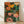 May include: A framed botanical art print leaning against a white wall on light wood flooring. The artwork features stylized mushrooms and flowers in orange, yellow, pink, and teal colors with dotted patterns and fern leaves on a dark background. Natural lighting creates soft shadows on the textured white fabric backdrop. The print is displayed in a simple light wood frame.