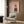 May include: Framed poster on wood-paneled wall showing a fluffy Pomeranian dog wearing olive green military uniform with beret and boots against pink background, with "DOGUE" text in orange letters at top. Modern beige armchair with patterned pillow, gold floor lamp, and dark side table with open book complete the contemporary room setting.