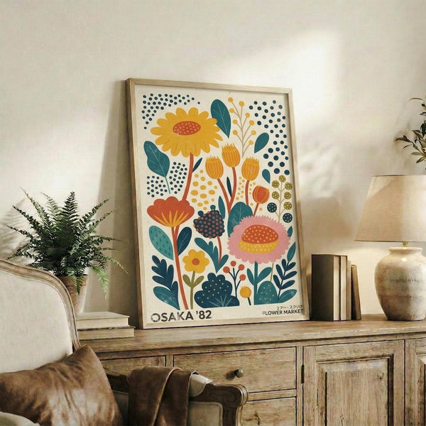 May include: Colorful botanical poster with stylized flowers in yellow, pink, orange, and teal displayed in light wood frame on rustic wooden sideboard. Scene shows cozy living room with cream chair, brown leather pillow, potted fern, ceramic vases, and stacked books. Artwork features folk art style blooms with polka dot patterns against cream background.
