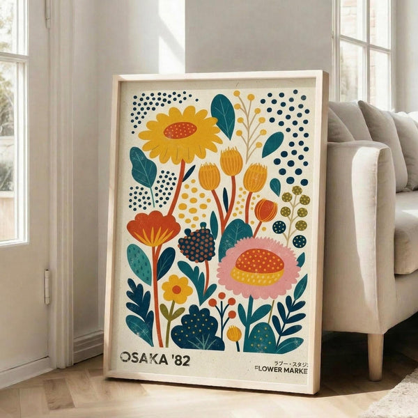 May include: Framed botanical poster print leaning against white wall next to beige sofa in bright room with windows. Artwork features colorful stylized flowers and plants in yellow, orange, pink, teal, and navy with decorative polka dot patterns on cream background. Text reads "Osaka '82 Flower Market" in black letters at bottom. Light wood frame, approximately 2-3 feet tall.