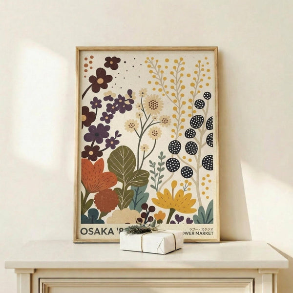 May include: Framed botanical poster with stylized flowers and plants in earth tones including purple, orange, yellow, and green against cream background, displayed on light wood shelf next to small wrapped gift with evergreen sprig. Artwork features abstract floral elements, leaves, and geometric patterns in mid-century modern illustration style within thin natural wood frame.