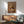 May include: Framed poster showing a cocker spaniel's head digitally placed on a human body wearing a brown blazer with flower lapel pin, styled as fashion magazine cover with "DOGUE" text at top. Displayed above wooden sideboard with white lamp, next to teal mid-century armchair in modern living room with white walls and natural lighting.