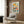 May include: Colorful floral poster print with stylized flowers in yellow, orange, pink, and teal displayed in light wood frame on brown wood panel wall above beige upholstered armchair with geometric throw pillow, alongside brass floor lamp and dark round side table with open book in modern living room setting