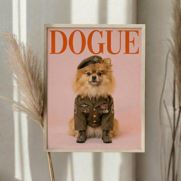 May include: Framed poster print displayed on white wall next to dried pampas grass stems. The artwork features a fluffy orange Pomeranian dog wearing a detailed olive green military uniform with medals, ribbons, boots and cap, set against a soft pink background. Bold orange "DOGUE" text appears at top in magazine-style lettering. Light wooden frame surrounds the print in bright, airy room setting.