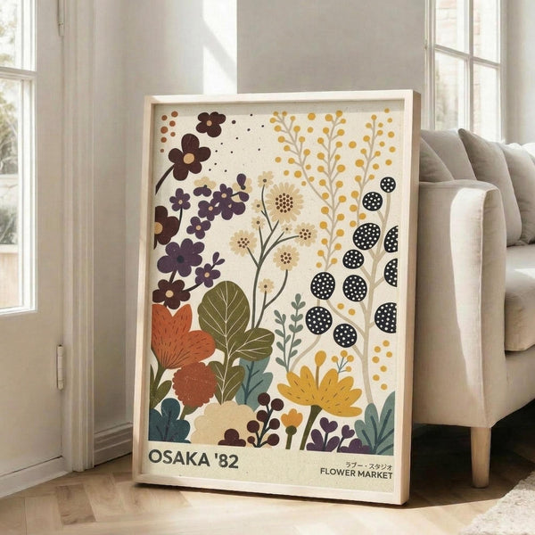 May include: Large framed botanical poster with stylized flowers in purple, orange, yellow, and teal colors against cream background, featuring text "OSAKA '82" and "FLOWER MARKET" at bottom. Artwork leans against white wall next to beige upholstered chair in bright, airy living room with tall windows and hardwood floors.