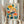 May include: Hands holding a framed botanical art print featuring colorful stylized flowers in yellow, orange, pink, and teal with dotted patterns and geometric leaves against a cream background. The print shows "OSAKA '82" and "FLOWER MARKET" text at bottom. Natural wood frame, pampas grass visible to left, soft neutral curtains in background creating bright, minimalist styling.