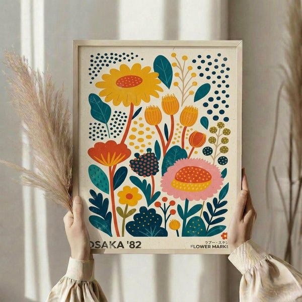 May include: Hands holding a framed botanical art print featuring colorful stylized flowers in yellow, orange, pink, and teal with dotted patterns and geometric leaves against a cream background. The print shows "OSAKA '82" and "FLOWER MARKET" text at bottom. Natural wood frame, pampas grass visible to left, soft neutral curtains in background creating bright, minimalist styling.