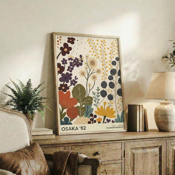 May include: Framed botanical poster with colorful illustrated flowers, leaves, and plants in purple, orange, yellow, and green tones displayed on rustic wooden sideboard. Scene shows cozy living room setting with beige armchair, fern plant, stacked books, ceramic vase, and neutral wall background. Natural lighting creates warm, inviting home decor atmosphere.