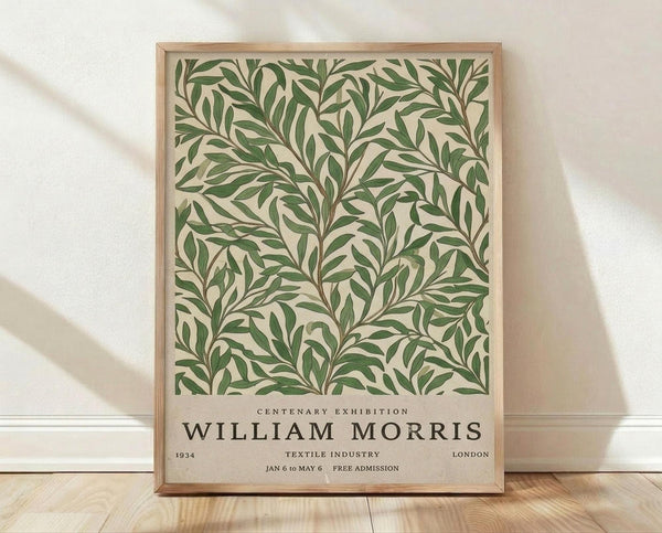 May include: Framed botanical poster with green leafy pattern on cream background, displayed against white textured wall on light wood floor. Text reads "Centenary Exhibition William Morris 1934 Textile Industry London Jan 6 to May 6 Free Admission." Natural wood frame, soft lighting creates diagonal shadows across scene.