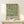 May include: Framed botanical poster with green leafy pattern on cream background, displayed on light wood furniture against white wall. Small wrapped gift box sits nearby. Natural lighting creates soft shadows. Frame appears to be light wood with clean lines.
