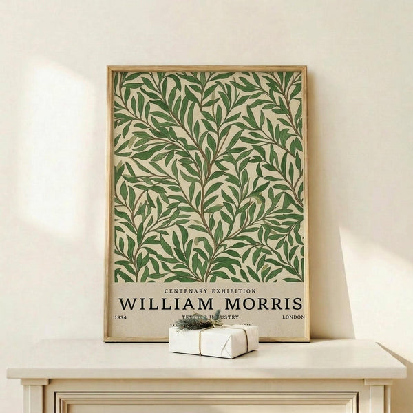 May include: Framed botanical poster with green leafy pattern on cream background, displayed on light wood furniture against white wall. Small wrapped gift box sits nearby. Natural lighting creates soft shadows. Frame appears to be light wood with clean lines.