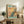 May include: Framed vintage-style poster featuring a blue and teal peacock perched on cherry blossom branches against a golden sunset sky, displayed on a rustic wooden sideboard next to potted plants, ceramic vases, and books in a cozy living room with neutral tones and natural lighting.