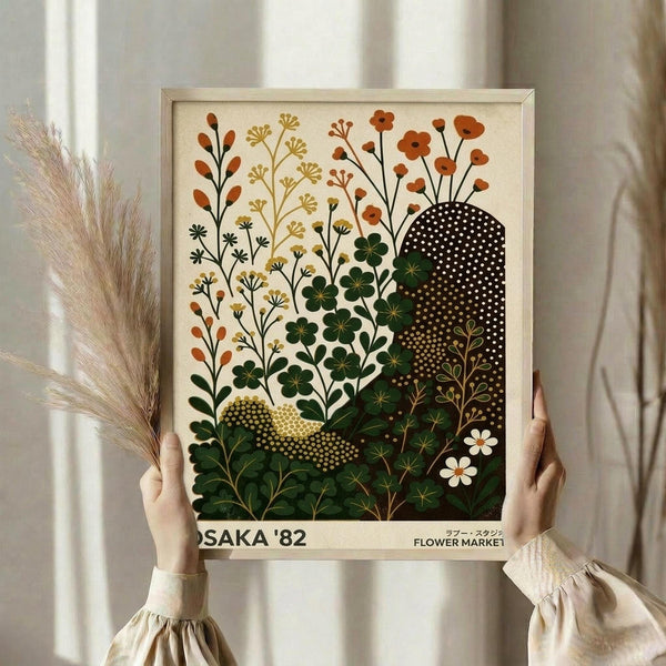 May include: Person in cream ruffled sleeves holding framed botanical art print featuring stylized wildflowers and plants in earthy tones of orange, green, and brown against cream background. Dried pampas grass visible to left, soft natural lighting on neutral curtained backdrop.