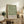 May include: Framed botanical poster with green leaf pattern on cream background displayed on rustic wooden sideboard beside fern plant, books, and ceramic vase in neutral-toned living room setting with beige chair visible