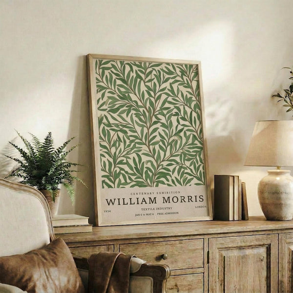May include: Framed botanical poster with green leaf pattern on cream background displayed on rustic wooden sideboard beside fern plant, books, and ceramic vase in neutral-toned living room setting with beige chair visible