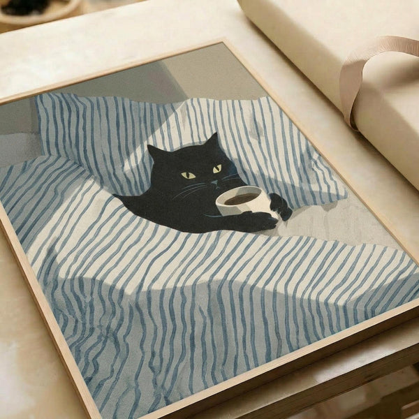 May include: Framed poster print displayed on light wooden coffee table next to cream-colored sofa. Artwork shows black cat with green eyes holding white coffee mug, lying on blue and white striped bedding or fabric with rumpled texture. Print has natural wood frame and is photographed in soft natural lighting.