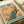 May include: Vintage-style poster print featuring a vibrant peacock perched on a cherry blossom branch against a golden sunset sky. The design includes "1967 Japan Week" text at the top and exhibition details at the bottom. The poster is shown lying flat on white paper with a white tube beside it, displaying traditional Japanese artistic style with rich blues, greens, and pink blossoms.