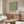 May include: Framed botanical poster with green leaf pattern and vintage exhibition text displayed above wooden sideboard in minimalist living room. White curved chair and glass coffee table in foreground. Poster features cream background with flowing green foliage design and black typography. Modern Scandinavian-style interior with natural wood tones and neutral colors.