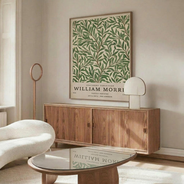 May include: Framed botanical poster with green leaf pattern and vintage exhibition text displayed above wooden sideboard in minimalist living room. White curved chair and glass coffee table in foreground. Poster features cream background with flowing green foliage design and black typography. Modern Scandinavian-style interior with natural wood tones and neutral colors.