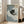 May include: Framed poster print leaning against white wall next to beige sofa in bright living room with large windows. Artwork shows black cat with yellow eyes holding white coffee mug, sitting on blue and white striped fabric or bedding. Print has light wood frame and soft, illustrated art style with muted colors.