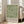 May include: Framed botanical poster with green leaf pattern on cream background, featuring exhibition text for William Morris 1934 Centenary Exhibition. Light wooden frame leans against white wall next to beige upholstered sofa in bright, naturally-lit room with white trim and windows.