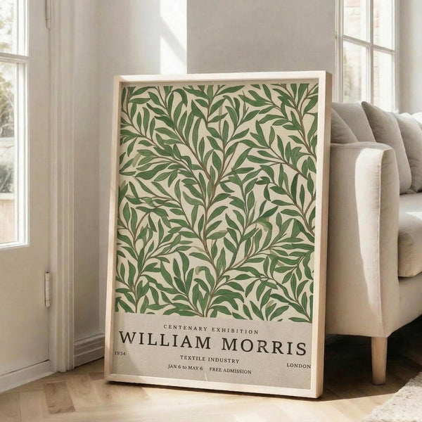 May include: Framed botanical poster with green leaf pattern on cream background, featuring exhibition text for William Morris 1934 Centenary Exhibition. Light wooden frame leans against white wall next to beige upholstered sofa in bright, naturally-lit room with white trim and windows.