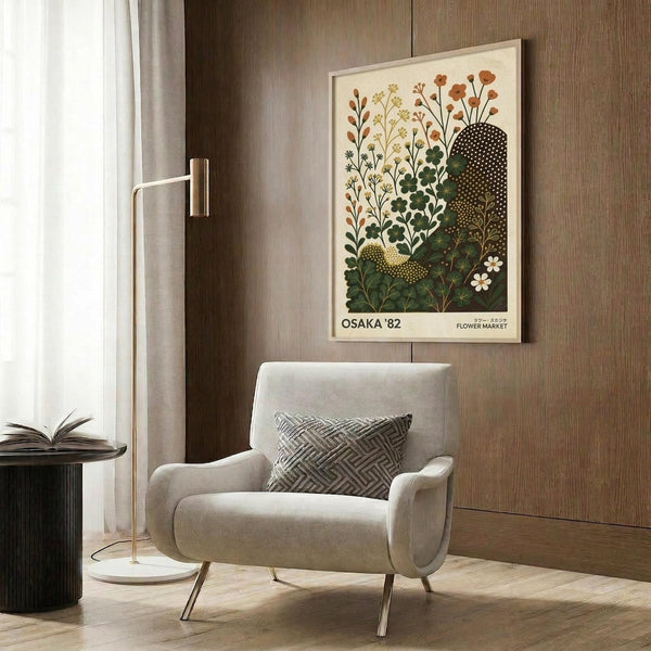 May include: Vintage botanical poster with stylized flowers and foliage in green, orange, and yellow tones, displayed in light wood frame on wood-paneled wall. Modern cream armchair with geometric patterned pillow sits below artwork. Brass floor lamp and dark round side table with open book complete the contemporary reading nook scene.