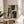 May include: Abstract geometric artwork in wooden frame featuring overlapping shapes in sage green, mustard yellow, cream, and charcoal colors, displayed on round wooden side table next to beige upholstered chair with white throw pillow, green plant leaves visible, soft natural lighting creating shadows on neutral wall