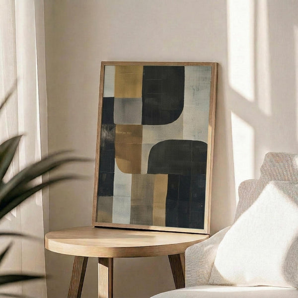 May include: Abstract geometric artwork in wooden frame featuring overlapping shapes in sage green, mustard yellow, cream, and charcoal colors, displayed on round wooden side table next to beige upholstered chair with white throw pillow, green plant leaves visible, soft natural lighting creating shadows on neutral wall