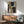 May include: Abstract geometric artwork with black, golden yellow, gray and cream colored shapes in wooden frame, displayed above mid-century modern walnut credenza with ceramic table lamp, next to sage green upholstered armchair with wooden arms in sunlit minimalist living room
