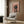 May include: Framed poster featuring a black and tan dachshund wearing a pink chef's hat and floral apron on coral pink background with "DOGUE" text at top, displayed above a cream upholstered armchair with geometric pillow in modern living room with wood paneled walls, brass floor lamp, and side table with open book.