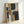 May include: Abstract geometric artwork in light wooden frame leaning against white wall. Features overlapping rectangular and curved shapes in muted palette of charcoal black, warm ochre yellow, soft grays, and cream. Painted with visible brushstrokes and textural elements creating layered, modern composition with organic flowing forms intersecting angular blocks.