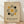 May include: Framed Bauhaus-style poster with geometric design featuring concentric rounded rectangles in black lines surrounding yellow and dark green circles on beige textured background, displayed against white wall on light wooden floor with natural lighting casting soft shadows