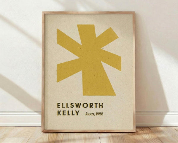 May include: Framed art print featuring a golden yellow abstract star or flower-like shape with eight pointed rays on cream textured background. Text below reads "ELLSWORTH KELLY" and "Aloes, 1958" in dark letters. Natural wood frame sits on light wooden floor against white wall with soft shadows from natural lighting.
