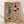 May include: Framed art print leaning against white wall on light wood floor. Illustration shows Mediterranean feast on coral pink background with blue vase of sunflowers, wine bottle and glass, decorative plate of whole fish with lemon slices and olives, bowl of pasta, fresh fruits including peaches and figs, and olive branches. Warm, hand-painted style with natural wood frame.