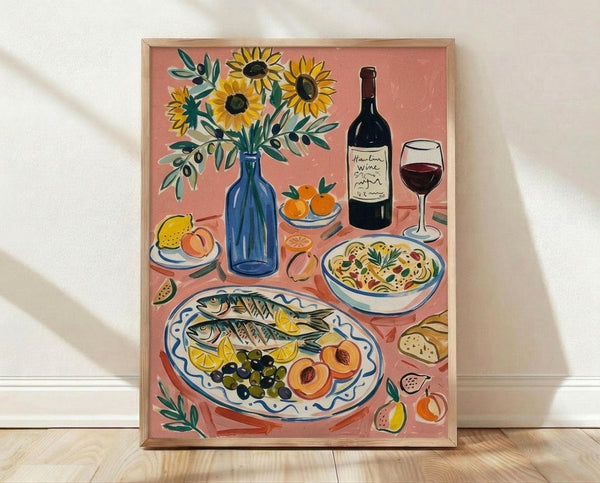 May include: Framed art print leaning against white wall on light wood floor. Illustration shows Mediterranean feast on coral pink background with blue vase of sunflowers, wine bottle and glass, decorative plate of whole fish with lemon slices and olives, bowl of pasta, fresh fruits including peaches and figs, and olive branches. Warm, hand-painted style with natural wood frame.