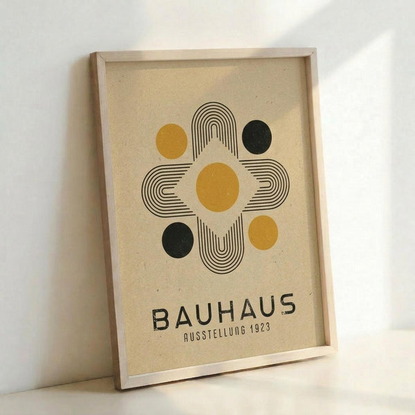 A framed poster featuring geometric Bauhaus-style design with concentric black line patterns forming a cross or flower shape. Four circles in mustard yellow and dark green are positioned around the central motif on cream-colored paper. The word "BAUHAUS" appears in bold black letters below with "AUSSTELLUNG 1923" in smaller text. The print is displayed in a light wooden frame leaning against a white wall.