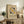 May include: Framed Bauhaus poster with geometric design featuring yellow and black circles arranged around striped linear elements on cream background, displayed on rustic wooden sideboard alongside books, ceramic vase, and fern plant in neutral-toned living room setting with beige armchair visible.