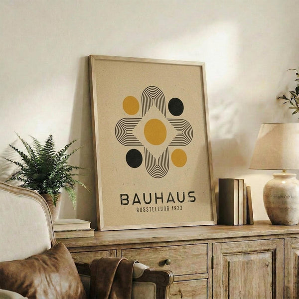 May include: Framed Bauhaus poster with geometric design featuring yellow and black circles arranged around striped linear elements on cream background, displayed on rustic wooden sideboard alongside books, ceramic vase, and fern plant in neutral-toned living room setting with beige armchair visible.