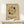 May include: Framed Bauhaus poster with geometric design featuring concentric rectangular lines forming a cross pattern, surrounded by mustard yellow and dark green circles on beige background. Natural wood frame displayed on light wooden shelf next to small white wrapped gift with twine and evergreen sprig against cream wall.