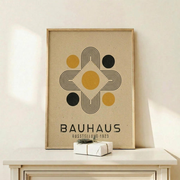 May include: Framed Bauhaus poster with geometric design featuring concentric rectangular lines forming a cross pattern, surrounded by mustard yellow and dark green circles on beige background. Natural wood frame displayed on light wooden shelf next to small white wrapped gift with twine and evergreen sprig against cream wall.