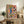 May include: Framed art print displayed on wooden sideboard in living room setting. Coral pink background with Mediterranean feast illustration featuring sunflowers in blue vase, wine bottle and glass, fish platter with olives and fruit, pasta bowl, and various produce. Styled with fern plant, books, ceramic vase, and cream chair visible in cozy home decor scene.