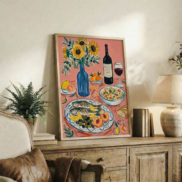May include: Framed art print displayed on wooden sideboard in living room setting. Coral pink background with Mediterranean feast illustration featuring sunflowers in blue vase, wine bottle and glass, fish platter with olives and fruit, pasta bowl, and various produce. Styled with fern plant, books, ceramic vase, and cream chair visible in cozy home decor scene.