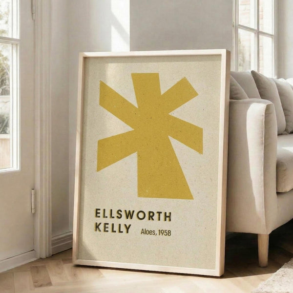May include: Framed poster featuring a yellow star-like abstract shape on cream textured background with "ELLSWORTH KELLY" and "Aloes, 1958" text below. Natural wood frame leaning against white wall beside beige upholstered sofa in bright living room with large windows and natural light streaming across wooden floors.