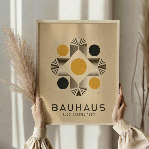 May include: Person in cream clothing holding a framed geometric poster with beige background, featuring abstract design of yellow and dark green circles arranged around curved black line patterns forming a cross-like shape, with "BAUHAUS" and "AUSSTELLUNG 1923" text below, while holding dried wheat stems, standing near white curtained window