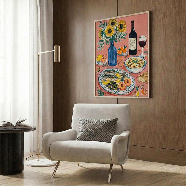 May include: A colorful illustrated poster print displayed in a modern living room setting with wood paneling. The artwork features a Mediterranean-style still life on coral pink background showing sunflowers in a blue vase, wine bottle and glass, fish on decorative plates, fruits, and olives. The framed print hangs above a cream upholstered armchair with geometric pillow, beside a brass floor lamp and dark round side table with open book.